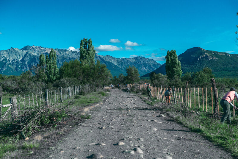 The image shows a rural road stretching into the distance, flanked by wooden fences and lush greenery. In the background, majestic mountains rise against a clear blue sky. Two people are working on the side of the road, adding a sense of human presence to the serene landscape. The scene evokes a sense of tranquility and connection with nature.